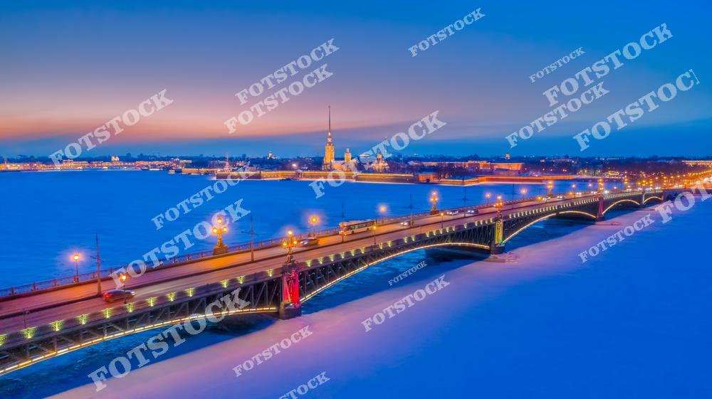 Фотография зимнего Санкт-Петербурга с высоты дрона, показывающая Петропавловскую крепость, реку Неву с ледяной поверхностью и мосты города в вечернее время. Снимок передает панорамный вид центра города, где историческая архитектура гармонично сочетается с зимними набережными и огнями вечернего Петербурга. Панорамный ракурс позволяет увидеть масштаб центральных улиц, величие крепости и динамику городской инфраструктуры, создавая атмосферу северной столицы России зимой. Изображение идеально подходит для туристических публикаций, зимних коллекций фотографий, постеров, интерьерного оформления и презентаций о городской архитектуре Петербурга.